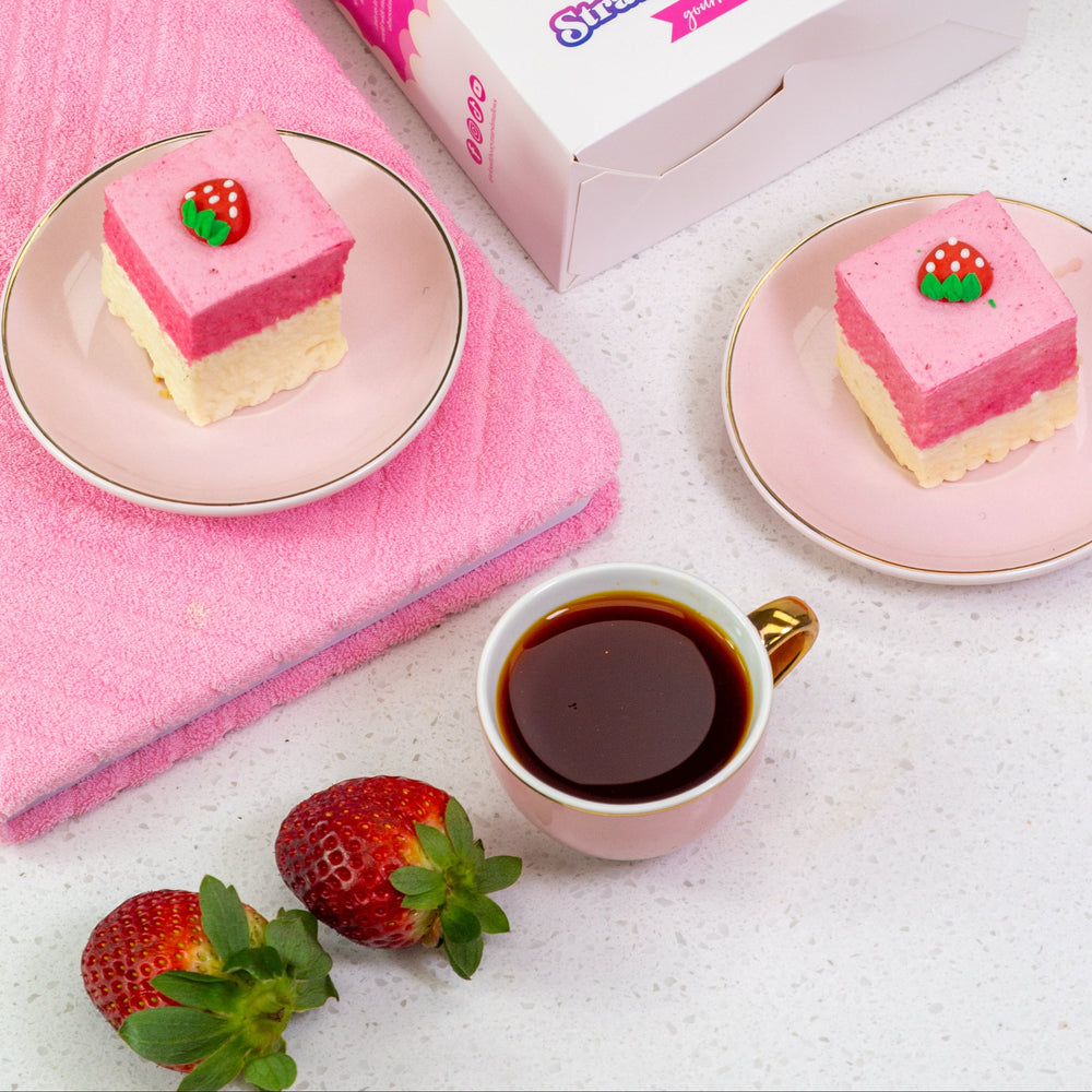 Pink dessert bars with strawberry decorations in a box, served on plates with a pink towel and cup of coffee.