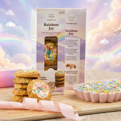 Cloud Theory Rainbow Joy cookies with packaging on a wooden board against a colorful, cloud-filled sky background.