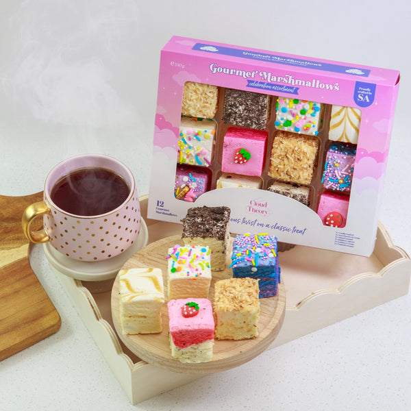 Colorful marshmallow treats on a wooden board with a cup of tea and a box of marshmallows in the background.