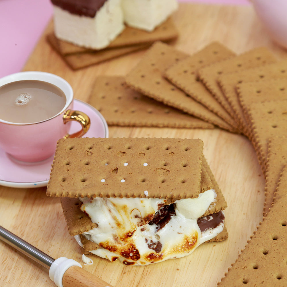 Golden honey-flavoured graham crackers stacked neatly next to marshmallows and chocolate, perfect for s’mores, cheesecake bases, and baking.