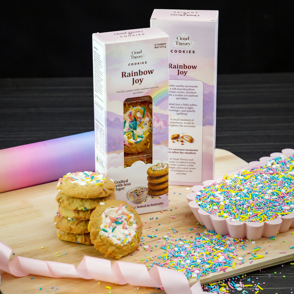 Rainbow Sprinkle with marshmallow cookies with colorful sprinkles on a wooden board, next to packaging for 'Rainbow Joy' cookies.