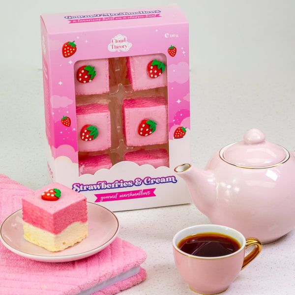 Pink cake with strawberry decoration, pink teapot, and cup of tea on a white surface.